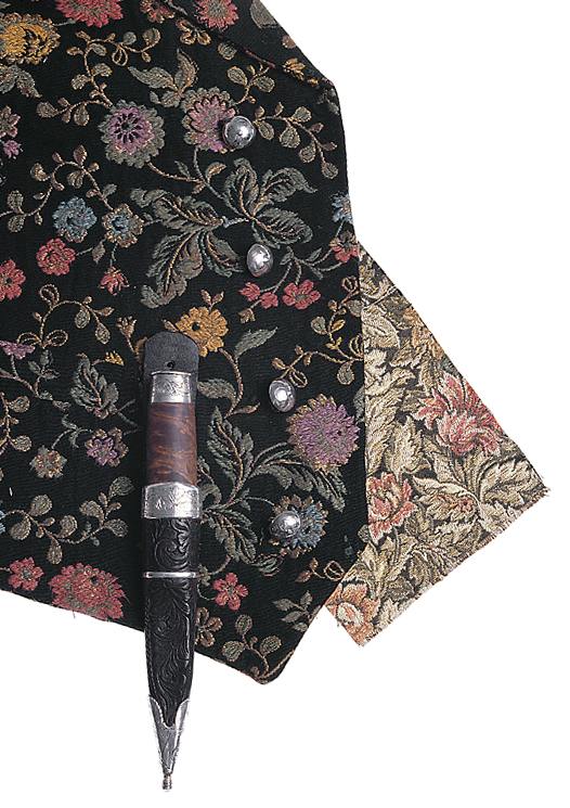 <p>Vesten er i småmønstret brokade eller svartvadmel. Ryggen er i grå lin. Den har oppstående krage og er dobbelspent med fem par sølvknapper.</p>